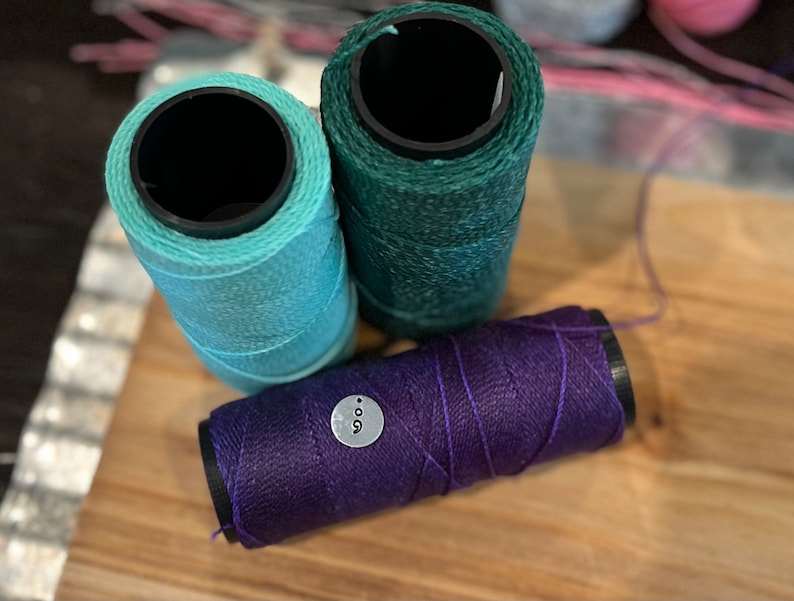May include: Three spools of thread in shades of teal, green, and purple. The purple spool has a small, round tag with the number 9 on it. The spools are arranged on a wooden surface, with a blurred background.