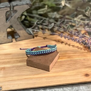 May include: A handmade friendship bracelet with a chevron pattern in teal and purple. The bracelet is displayed on a small brown block, set against a wooden background with a metal letter "K" and dried lavender.