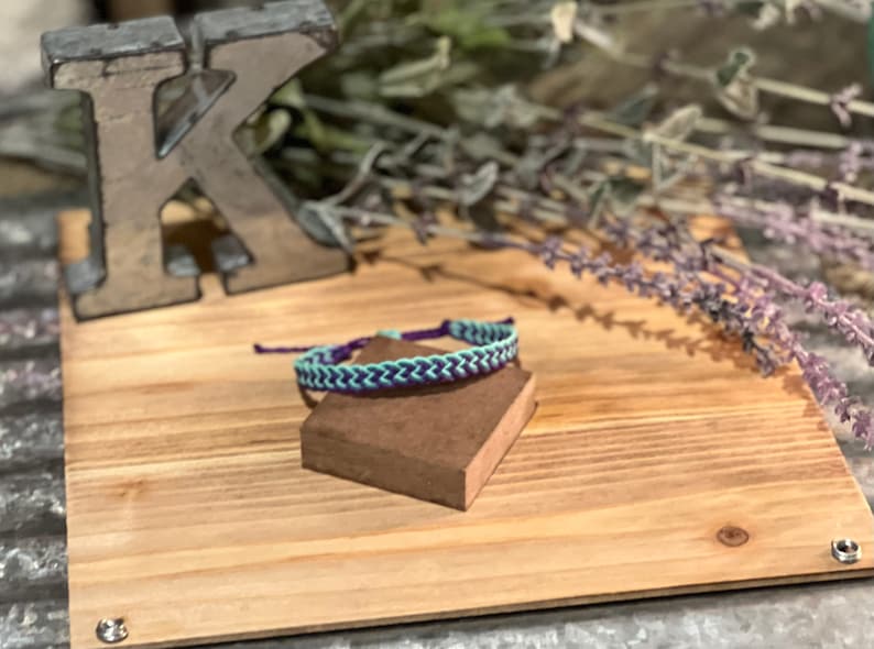 May include: A handmade friendship bracelet with a chevron pattern in teal and purple. The bracelet is displayed on a small brown block, set against a wooden background with a metal letter "K" and dried lavender.