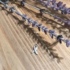 May include: A small, silver-colored ribbon charm, a symbol of awareness, rests on a wooden surface. Lavender sprigs are arranged in the background, casting shadows. The charm is a simple, elegant design.