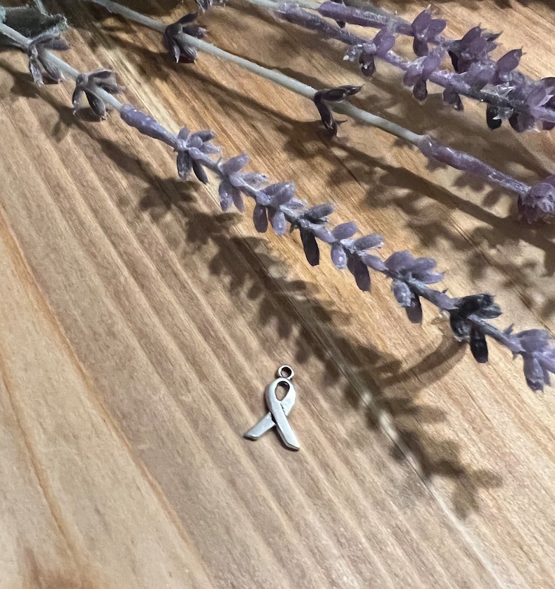 May include: A small, silver-colored ribbon charm, a symbol of awareness, rests on a wooden surface. Lavender sprigs are arranged in the background, casting shadows. The charm is a simple, elegant design.