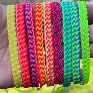 Może przedstawiać: Kolekcja kolorowych, plecionych bransoletek w różnych neonowych kolorach, w tym żółtym, różowym, pomarańczowym, fioletowym, niebieskim i zielonym. Każda bransoletka wykonana jest z tkanych nici i posiada zapięcie na pętelkę. Bransoletki są prezentowane na rozmytym zielonym tle.