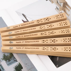 K&ouml;nnte beinhalten: Ein Stapel von f&uuml;nf Holzf&auml;cher mit filigranen Ausschnitten. Jeder F&auml;cher ist mit dem Text "Sophia & Michael 12.20.2023" personalisiert.