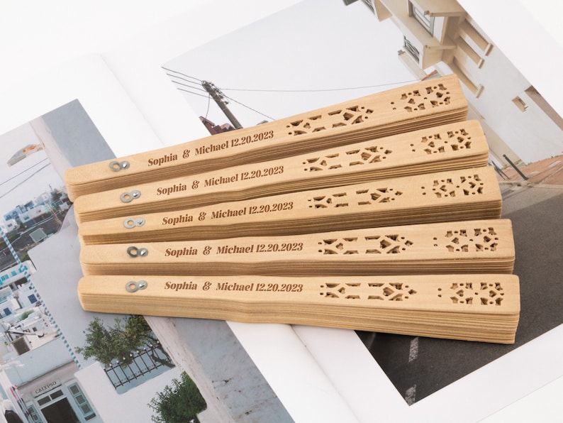 K&ouml;nnte beinhalten: Ein Stapel von f&uuml;nf Holzf&auml;cher mit filigranen Ausschnitten. Jeder F&auml;cher ist mit dem Text "Sophia & Michael 12.20.2023" personalisiert.