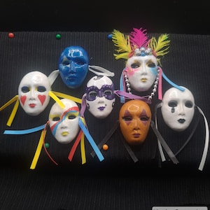 May include: A collection of seven small decorative face masks featuring various painted designs, including a blue mask with stars, a white mask with red hearts, a white mask with colorful stripes, a white mask with purple accents, and a white mask with a teardrop.
