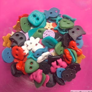 Puede incluir: Una colección de pequeños botones variados en diversas formas, incluyendo peces, estrellas y flores, con una paleta vibrante de rojo, azul, verde, negro, blanco y rosa. Los botones están esparcidos, mostrando diferentes texturas.