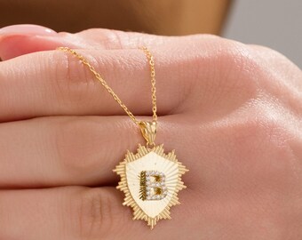 Puede incluir: Collar dorado con un colgante con la letra "B" incrustada con pequeñas piedras transparentes. El colgante tiene forma de escudo con un diseño de rayos de sol. La cadena es delicada.
