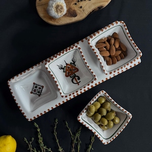 May include: A white ceramic serving dish with three compartments. The dish has a brown and white polka dot pattern and features decorative designs in each compartment. The dish is filled with almonds, olives, and a decorative design.