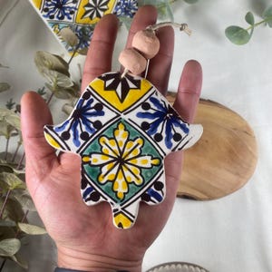 May include: A ceramic hamsa hand with a blue, green, yellow and white geometric pattern. The hamsa is decorated with a floral design and has a string with two small pink beads attached.