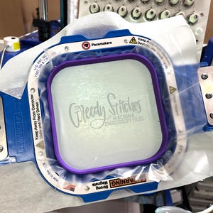 May include: An embroidery machine hoop with a purple inner frame and a blue outer frame. The text "Greedy Stitches MACHINE EMBROIDERY FILES" is visible on the fabric. The machine has multiple needles and the text "WARNING Strong Magnets" is also visible.
