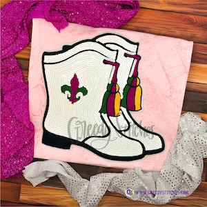 May include: A pair of white cowboy boots with black trim, embellished with small, sparkling accents. The boots feature a fleur-de-lis and colorful tassels. The design is set against a pink background, with sequined fabric accents.