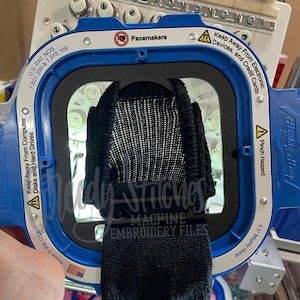 May include: A black knitted item is held within a blue embroidery hoop. The hoop has safety warnings and the text "Mighty Hoop" and "Machine Embroidery Files". The image also shows parts of an embroidery machine.