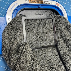 May include: A blue 5.5 inch Mighty Hoop embroidery frame with strong magnets, holding a grey heather knit beanie. A black plastic S.S Beanie embroidery jig is positioned inside the hoop, securing the fabric for machine embroidery work.