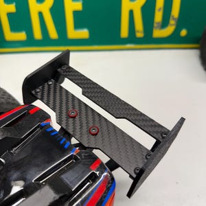 May include: Black and carbon fiber RC car wing with red accents. The wing is attached to a black and multi-colored RC car body. The background includes a green sign with yellow lettering that reads "ERE RD."