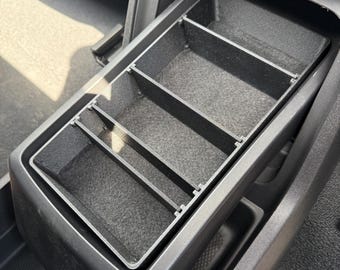 May include: A black plastic center console organizer insert featuring four rectangular compartments with a felt-lined base. The organizer is designed to fit into the deep storage well of a vehicle console, providing segmented storage space.