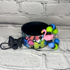 May include: Black drink holder with a colorful tropical design featuring pink flamingos and green bows. A black bow-shaped accessory and a beaded bracelet with pink flamingo and colorful beads are also present.