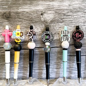May include: Eight colorful pens with different religious themed charms and beads. The charms include crosses, sunflowers, and inspirational words like "Fear Not", "Faith Over Fear", and "Pray".