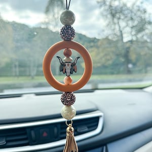 May include: Car hanging ornament featuring a circular orange ring with a bull skull design. The ornament includes decorative beads in shades of brown, cream, and gold, and a tassel. The ornament hangs from a black string.