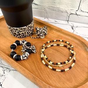 May include: A black tumbler with a leopard print sleeve sits on a wooden tray, alongside a matching bow and two beaded bracelets. One bracelet is black with silver accents, and the other is a stack of three beaded bracelets in neutral tones.