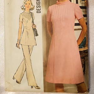 May include: Vintage sewing pattern featuring two designs: a light pink dress with short sleeves and a high neckline, and a cream-colored pantsuit with a short-sleeved top. The pattern includes the word "DESIGN" vertically on the side.