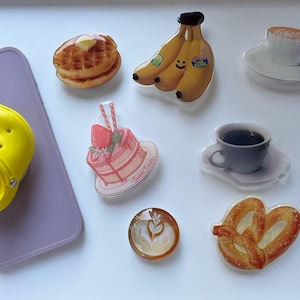May include: A collection of food-themed acrylic charms, including a waffle with butter, a bunch of bananas, a slice of cake, coffee cups, a pretzel, and a latte. A yellow miniature shoe is attached to a purple phone case.