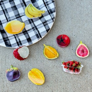 May include: A selection of fruit stickers, featuring a bunch of bananas, a lemon, a lychee, a tomato, a fig, a mangosteen, a carambola, a half-cut orange, and a punnet of strawberries. The stickers are on a checkered plate and a grey surface.