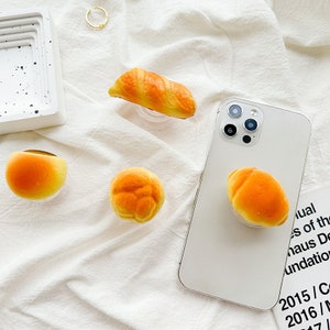 May include: Four different phone grips shaped like various types of bread. The grips are made of a soft, squishy material and are designed to be attached to the back of a phone.