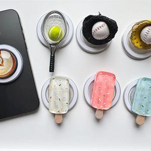 May include: Assortment of phone grips with various designs, including a coffee cup, tennis racket, baseball glove, and popsicles. The phone grips are white with a clear plastic top and a black base.