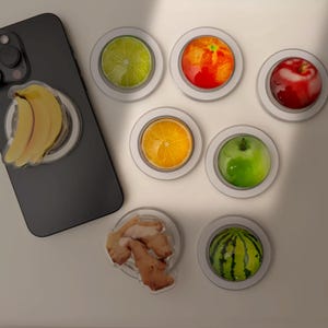 Puede incluir: Una colección de agarres para teléfono con temática de frutas, que incluyen un plátano, lima, naranja, manzana, jengibre y sandía. Los agarres son redondos con un acabado transparente y brillante y bordes blancos. También se ve un teléfono inteligente negro.