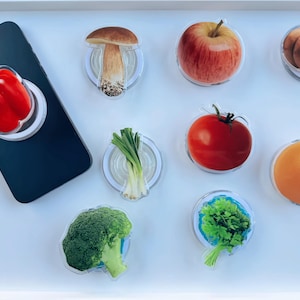 以下が含まれることがあります： 赤ピーマン、青ネギ、トマト、キノコ、ブロッコリー、パセリ、オレンジ、リンゴ、カボチャなど、さまざまな果物や野菜をモチーフにしたカラフルなスマホグリップのセットです。