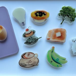 Pode incluir: Uma coleção de autocolantes decorativos com temas de comida e outros, incluindo uma lâmpada, um ovo, uma sobremesa, torradas com manteiga, bananas, cartas de jogar e uma chávena de café. Uma capa de telemóvel roxa também está presente.