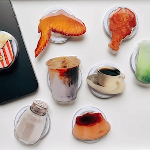 May include: Assortment of phone grips featuring various food and drink designs.  The designs include popcorn, fried chicken, coffee, a green drink, a salt shaker, and a dessert.
