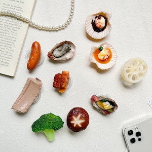 May include: A collection of miniature food items on a white surface. The items include a sausage, an oyster, a piece of meat, a broccoli floret, a mushroom, a scallop, a lotus root, and a phone case with a camera.