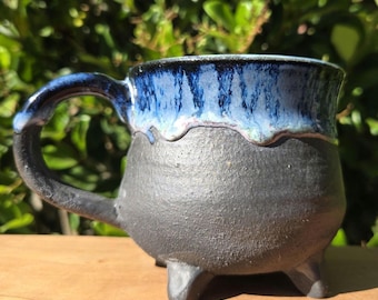 May include: A handmade ceramic mug with a unique design. The mug features a dark gray base and a blue glazed rim. It has a handle and three small feet. The blue glaze drips down the sides, creating a textured effect. The mug is set on a wooden surface.