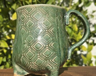 May include: A sage green ceramic mug featuring a raised diamond pattern across its exterior surface. The mug has a curved handle on the right side and stands on three small, integrated feet at the base, showcasing a glossy, textured finish.