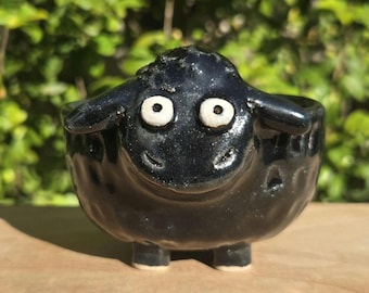 May include: A small, glossy black ceramic bowl shaped like a sheep. The bowl features a textured, rounded body, two small feet, and a face with two wide, white-rimmed eyes and a simple carved smile. The surface has a reflective, dark glaze finish.