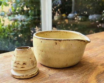 May include: A light yellow ceramic bowl with a small pouring spout and a matching conical ceramic stand. Both pieces feature a speckled texture with dark brown flecks and a smooth, matte finish, showcasing a handcrafted appearance.
