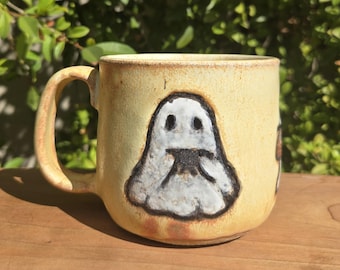 May include: A ceramic mug with a light yellow glaze and a ghost design. The ghost is white with black details. The mug has a rounded shape and a large handle. The mug is sitting on a wooden surface.