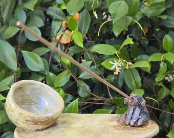 May include: A handmade ceramic incense holder featuring a flat, oval-shaped base with a smooth, light beige finish. On one side sits a small, deep bowl, and on the opposite side is a dark, textured figurine of a dog holding a thin incense stick.