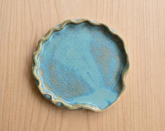 May include: A handmade ceramic plate with a wavy edge. The plate has a turquoise glaze with darker blue and brown accents. The plate is round and sits on a light wood surface.