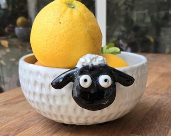 May include: A white ceramic bowl with a textured exterior, featuring a black and white sheep face on the front. The bowl is filled with bright yellow lemons. The bowl is sitting on a wooden surface.