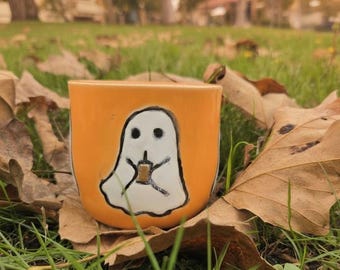 May include: Orange ceramic mug featuring a white ghost illustration holding a beverage. The ghost has two black dot eyes and a black mouth. The mug is sitting on a bed of grass and fallen leaves.