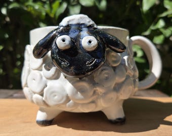 May include: A white ceramic mug shaped like a sheep with a glossy black face, protruding round eyes, and a textured body featuring raised spiral patterns. The mug has a curved handle on the right side and stands on three short, sturdy white legs.