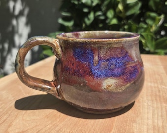 May include: A ceramic mug with a rounded body and a curved handle. The exterior features a reactive glaze with blended tones of deep purple, vibrant blue, and earthy brown, creating a mottled, glossy finish across the surface of the vessel.