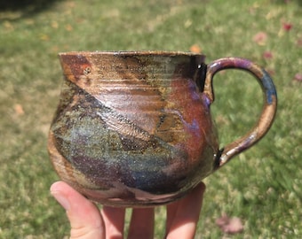May include: A ceramic mug with a rounded body and a curved handle, featuring a variegated glaze in shades of metallic bronze, deep brown, dark blue, and hints of purple, with a glossy finish and visible texture across the entire exterior surface.