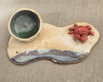 May include: A ceramic tray featuring a small, round, deep bowl with a green and blue interior glaze. The tray base is beige with a textured blue and purple edge, and a small, sculpted, bright red crab is attached to the surface of the tray.