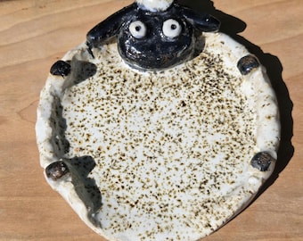 May include: A ceramic sheep-shaped dish with a black face and ears, white eyes, and a white and speckled body. The dish has a rounded shape with black accents on the edges. The background is a wooden surface.