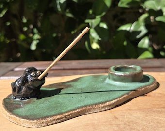 May include: A ceramic incense holder with a green glazed surface and a dark brown, textured base. The piece features a small, dark, sculpted frog figure on one end holding a thin incense stick, and a circular indentation on the other end.