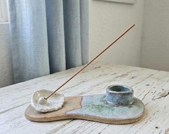 May include: A ceramic incense holder with a smooth, elongated base featuring a speckled tan and seafoam green glaze. The piece includes a small, white, shell-shaped dish for holding an incense stick and a raised, cylindrical cup for a candle.