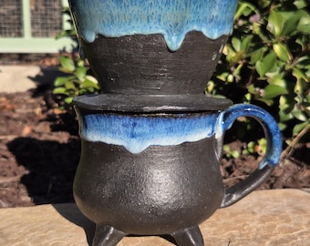 May include: A handmade ceramic coffee pour-over set. The set includes a black mug with three small feet, a matching lid, and a pour-over cone. The top and rim of the mug and cone are glazed in a vibrant blue.
